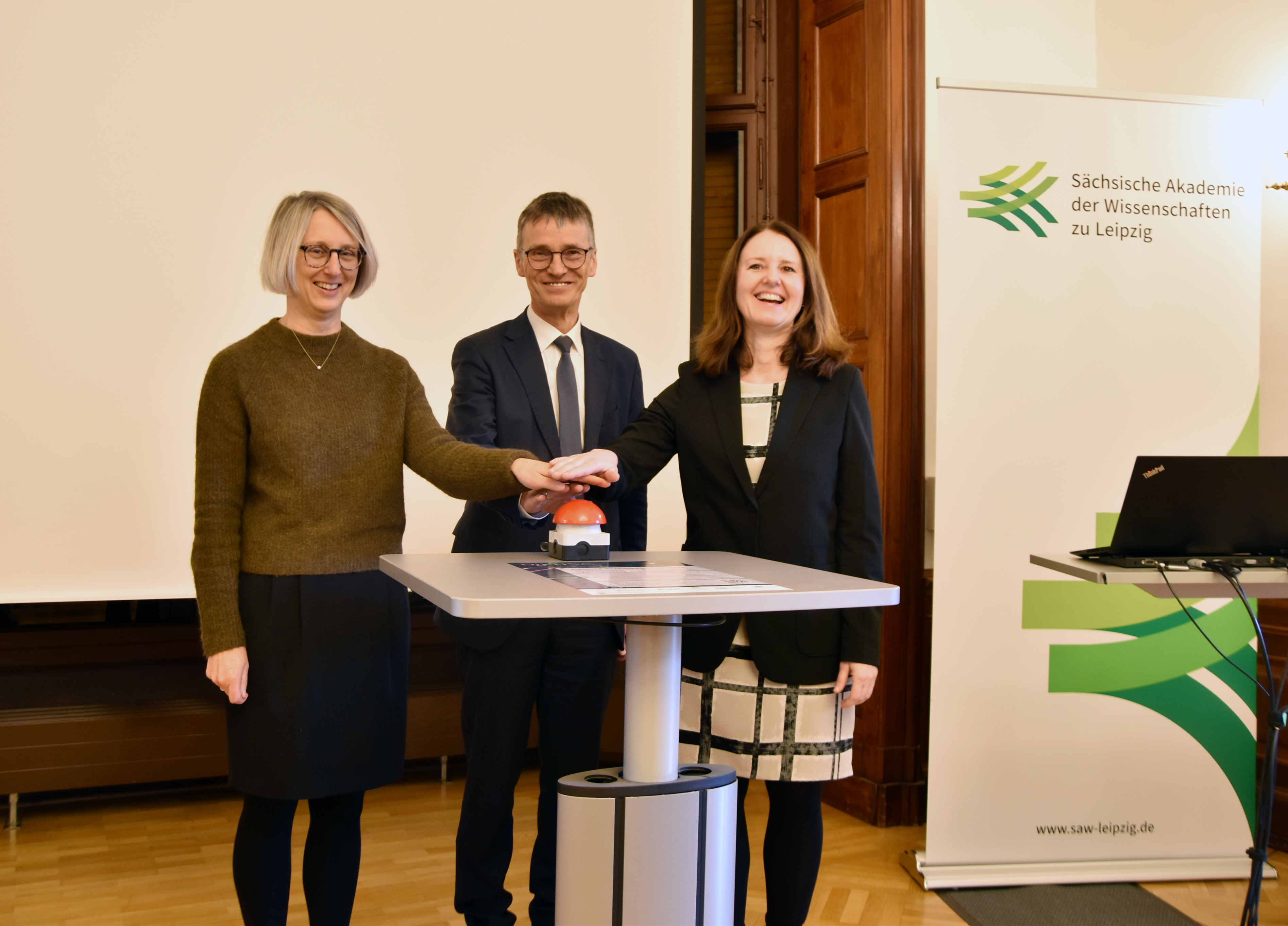 An einem Stehtisch stehen zwei weibliche und eine männliche Personen und betätigen zu dritt einen roten Knopf. Die Person rechts im Bild (Staatssekretärin Graßmann) lacht, die anderen beiden lächeln. Im Hintergrund sieht man einen Aufsteller mit dem Logo der Sächsischen Akademie der Wissenschaften.