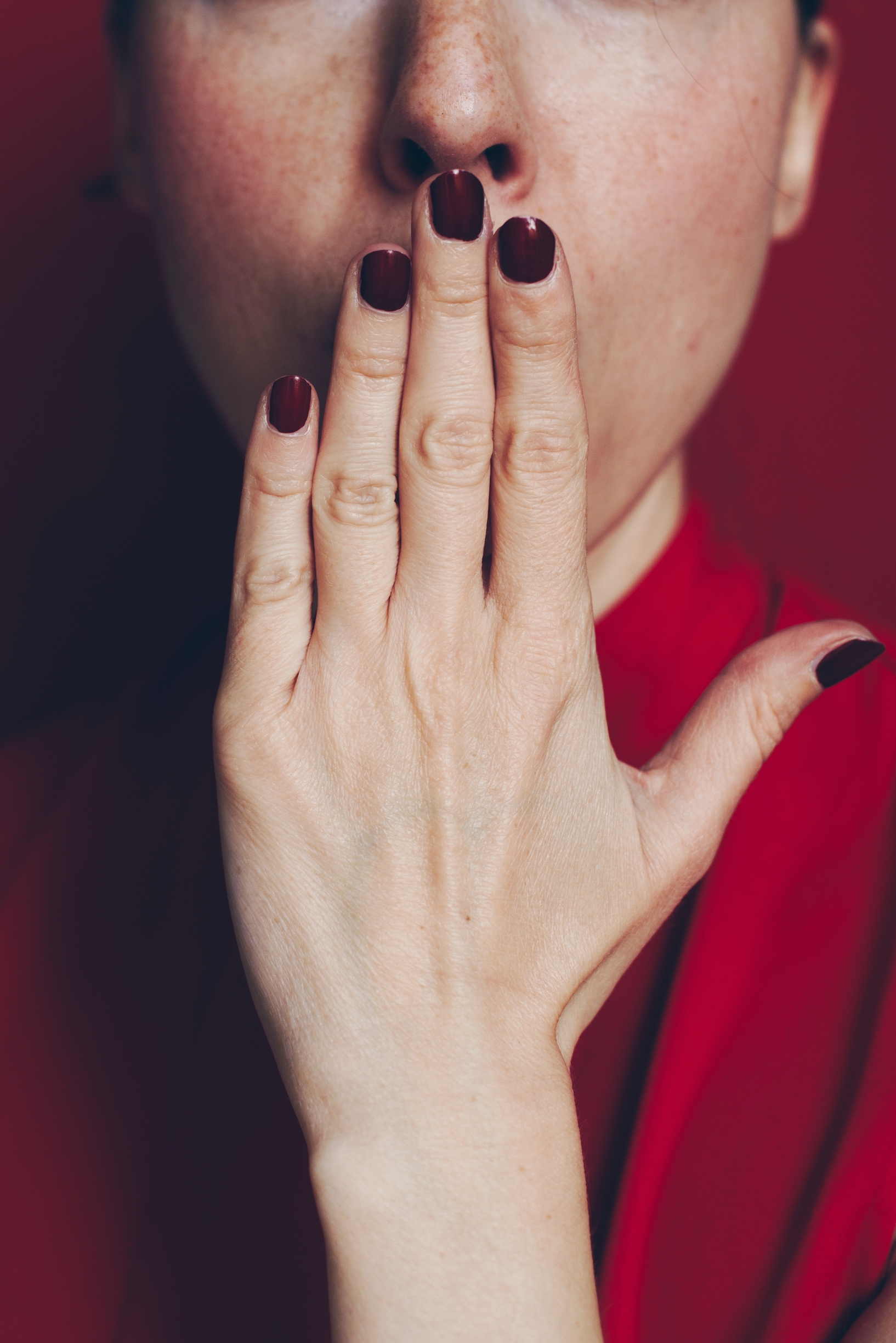 Ausschnitt eines weiblichen Gesichtes ab Nase abwärts vor rotem Hintergrund. Die Frau legt sich ihre Hand mit tiefroten Fingernägeln über den Mund.