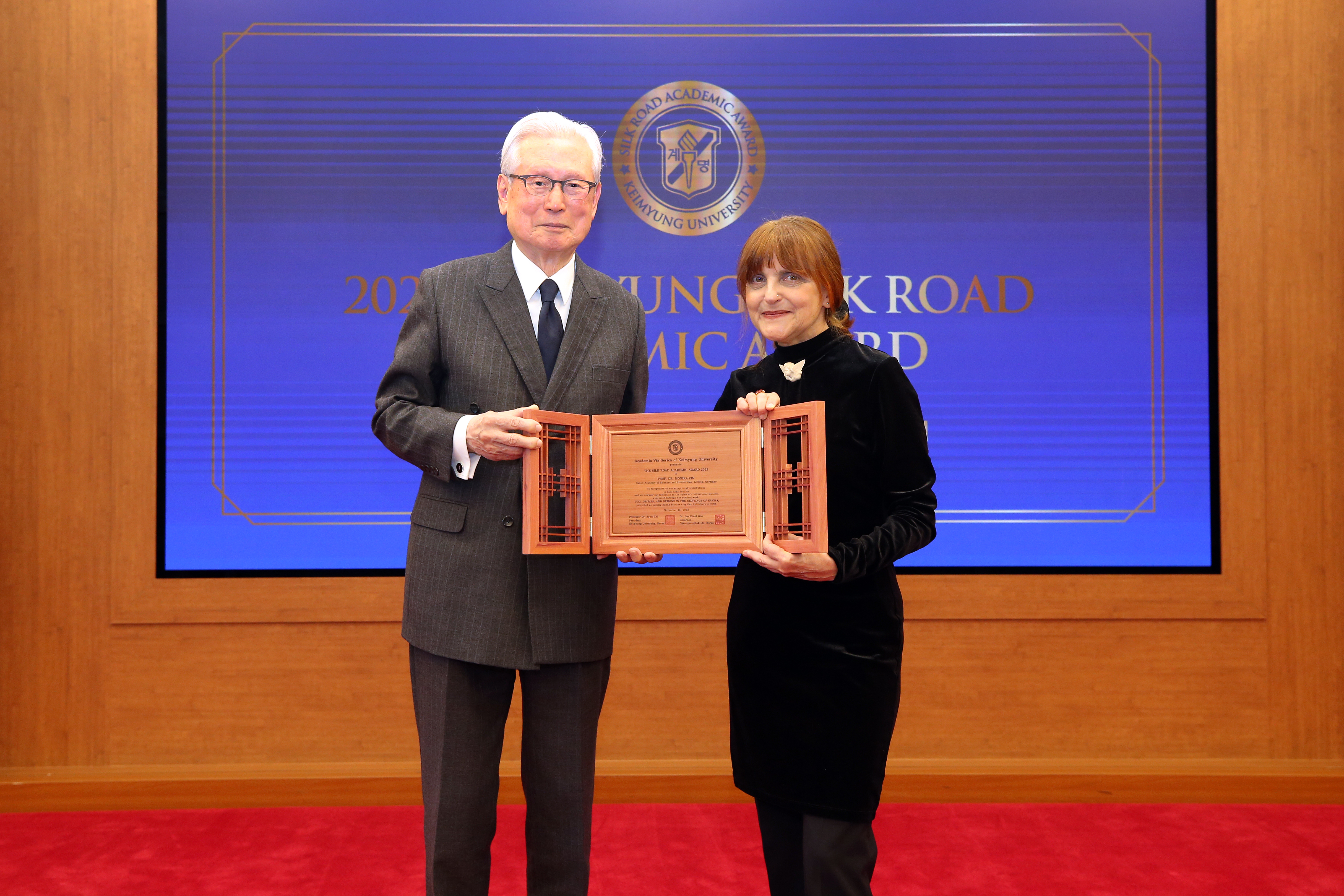 Prof. Monika Zin is presented with the award which consist of three pieces of wood in front of a dark blue screen. 