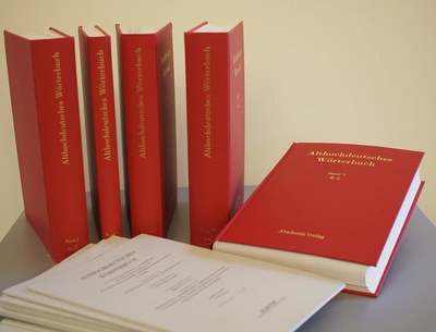 Vier rote Wörterbuchbände stehen auf einem Tisch, daneben liegt ein roter Band. Davor liegen Papierbündel mit veröffentlichen Teilen des Wörterbuchs.